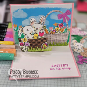 Handmade pop-up Easter card featuring two white bunnies, colorful eggs, and spring flowers in front of a large basket under a blue sky. “Easter's on its way” is written inside. Markers and craft supplies surround the card. To read more, visit Patty Stamps (pattystamps.com)