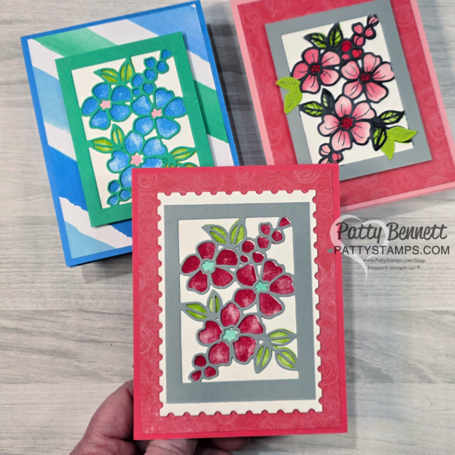 Three handmade greeting cards are displayed on a gray wood surface. Each features a layered rectangle with colorful die-cut floral designs and green leaves. The cards have blue, pink, and red color themes and include a credit watermark “Patty Bennett Pattystamps.com.”. To read more, visit Patty Stamps (pattystamps.com)