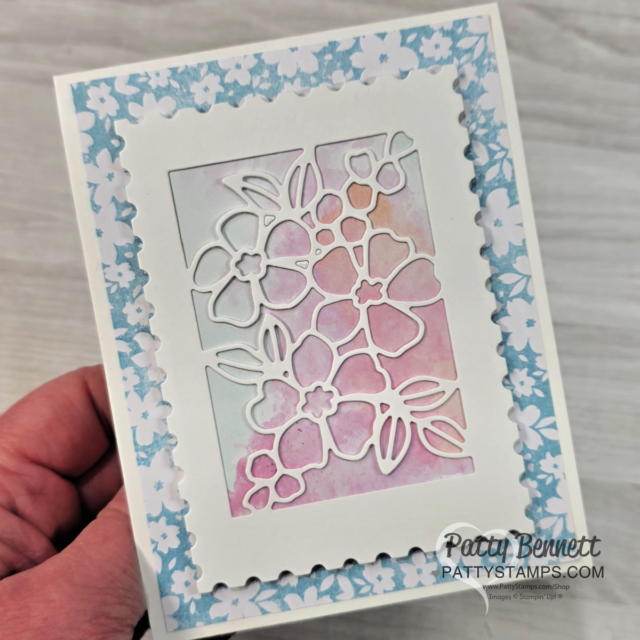 A hand holds a handmade greeting card with a blue floral border, scalloped white frame, and a die-cut overlay of flowers revealing a pastel pink, blue, and peach watercolor background. "Patty Bennett" and "PATTYSTAMPS.COM" are printed in the bottom right corner. To read more, visit Patty Stamps (pattystamps.com)