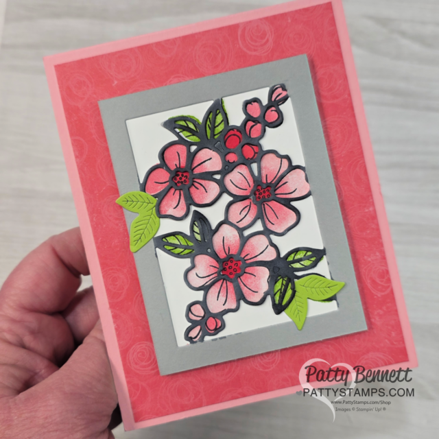 A hand holds a handmade greeting card with pink and coral flower cutouts, green leaves, and red berries framed by gray and white rectangles. The card has a coral pink background with subtle floral patterns. "Patty Bennett" and "PattyStamps.com" are printed at the bottom. To read more, visit Patty Stamps (pattystamps.com)