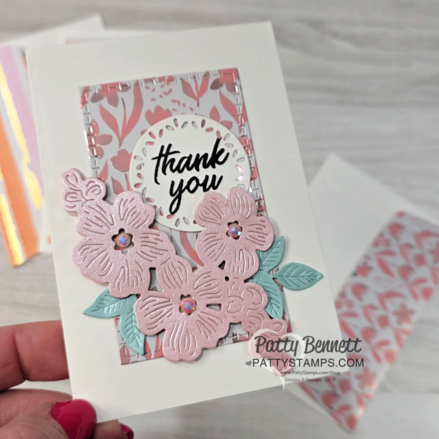 A hand holds a white card featuring layered pink and blue die-cut flowers with gem centers, green leaves, and a lace-edged circle with “thank you” in script. Behind, a pink patterned rectangle adds depth. Two more similar cards sit on a light surface in the background. To read more, visit Patty Stamps (pattystamps.com)
