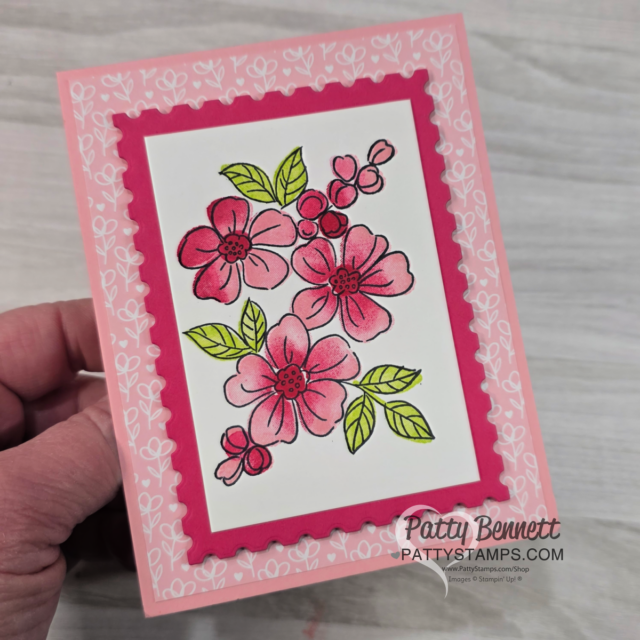 A hand holds a pink floral greeting card with a white center, featuring hand-colored pink flowers and green leaves. The card has a dark pink scalloped frame, light pink border, and “Patty Bennett PATTYSTAMPS.com” printed at the bottom right. To read more, visit Patty Stamps (pattystamps.com)