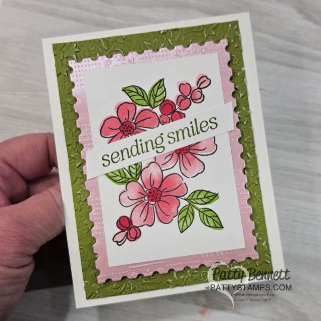 A hand holds a handmade card featuring pink and red flowers with green leaves. The card has a light pink border, an embossed olive-green frame, and a banner that reads "sending smiles." “Patty Bennett” and “PattyStamps.com” are printed in the corner. To read more, visit Patty Stamps (pattystamps.com)