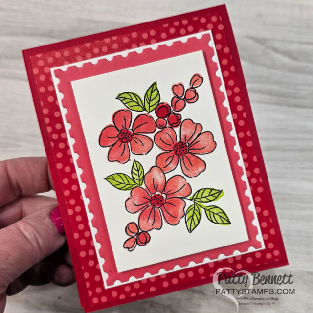 A handmade card featuring red and pink flowers with green leaves, bordered by a white scalloped frame and set against a red polka dot background. A hand holds the card. “Patty Bennett pattystamps.com” is printed at the bottom right corner. To read more, visit Patty Stamps (pattystamps.com)