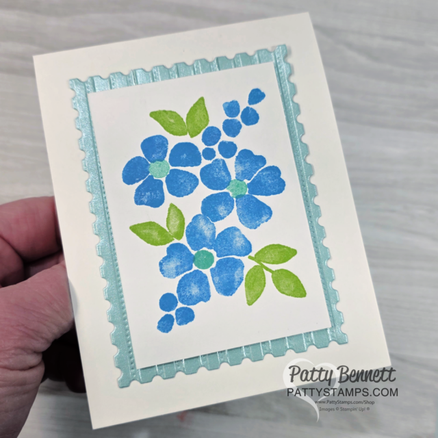 A hand holds a handmade card with a blue and green floral design. The flowers and leaves are stamped on a white rectangle, framed by a light blue border with a scalloped edge. “Patty Bennett PATTYSTAMPS.COM” is printed in the bottom right corner. To read more, visit Patty Stamps (pattystamps.com)