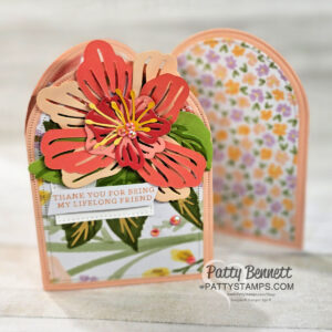 A handmade, arched greeting card with layered pink, red, and yellow flower embellishments and green leaves on the front. A small tag reads “Thank you for being my lifelong friend.” The background shows floral patterns. “Patty Bennett pattystamps.com” is watermarked. To read more, visit Patty Stamps (pattystamps.com)