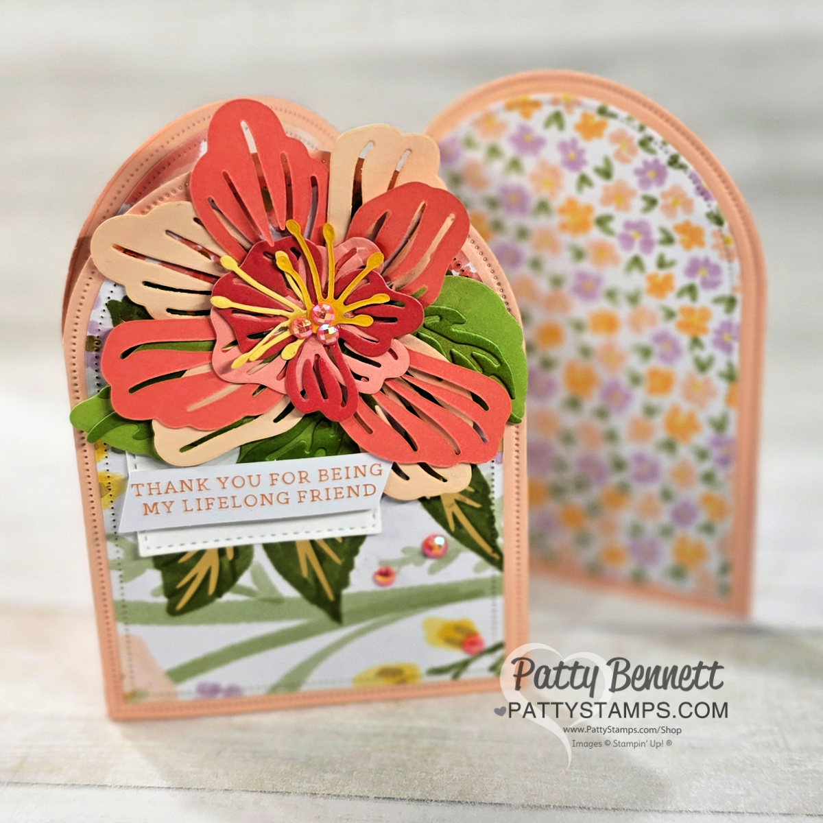 A handmade, arched greeting card with layered pink, red, and yellow flower embellishments and green leaves on the front. A small tag reads “Thank you for being my lifelong friend.” The background shows floral patterns. “Patty Bennett pattystamps.com” is watermarked. To read more, visit Patty Stamps (pattystamps.com)