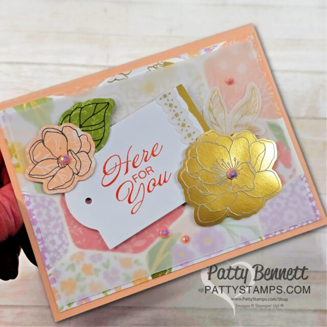 A handmade greeting card featuring layered pastel flowers, gold foil floral accents, green leaves, and a tag with “Here for you” in coral script. The background has a soft floral pattern, and three small pearl embellishments are added for detail. To read more, visit Patty Stamps (pattystamps.com)