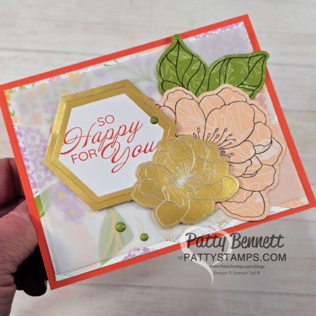 A hand holds a colorful, handmade greeting card featuring layered paper flowers in gold and peach, green leaves, and a hexagonal panel with red text reading “So Happy For You.” The card background is soft floral, and “Patty Bennett PattyStamps.com” is printed at the bottom. To read more, visit Patty Stamps (pattystamps.com)