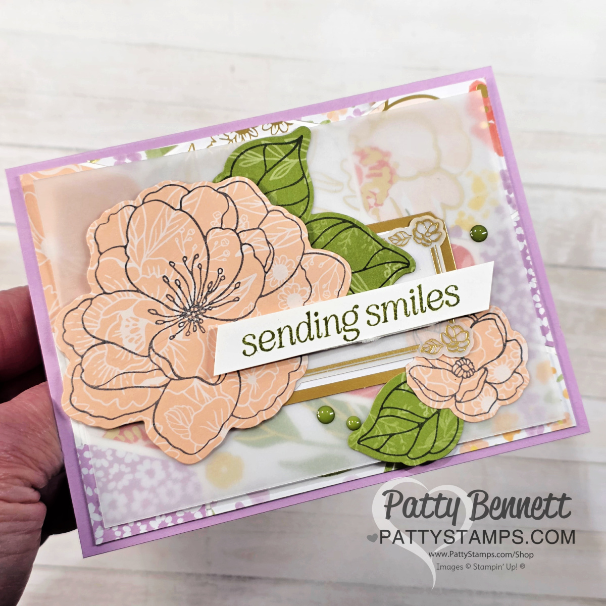 A hand holds a layered greeting card with peach-colored, outlined flowers, green leaves, and the message "sending smiles" on a white strip. The background has soft floral patterns and green dots. "Patty Bennett pattystamps.com" is watermarked on the card. To read more, visit Patty Stamps (pattystamps.com)