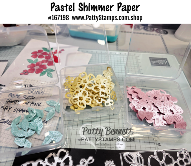 A close-up of three plastic containers with pastel paper die-cut floral shapes in blue, yellow, and pink. The containers sit on a craft mat beside a stamped flower image. Text at the top reads "Pastel Shimmer Paper #167198 www.PattyStamps.com.shop". To read more, visit Patty Stamps (pattystamps.com)