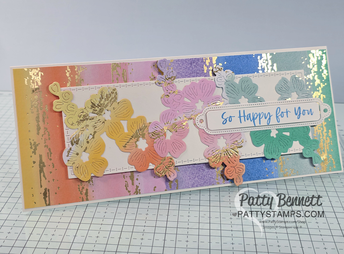 A handmade card features pastel rainbow stripes with gold foil accents as the background. Layered die-cut butterflies in pastel shades and a stitched white rectangle decorate the front. A white sentiment label reads “So Happy for You.” The design sits on a grid mat. To read more, visit Patty Stamps (pattystamps.com)