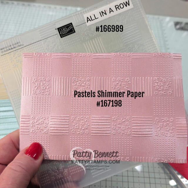 A hand with red nail polish holds embossed pink paper labeled “Pastels Shimmer Paper #167198” in front of a clear embossing folder labeled “ALL IN A ROW #166989.” Both items are shown on a craft table with grid lines in the background. To read more, visit Patty Stamps (pattystamps.com)