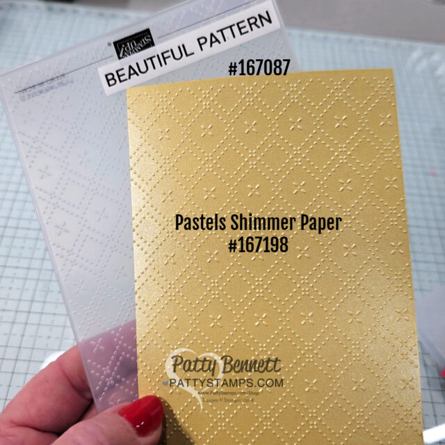 A hand with red nail polish holds a sheet of yellow pastels shimmer paper embossed with a diamond pattern. Behind it, a clear plastic embossing folder labeled "BEAUTIFUL PATTERN #167087" is visible. The background shows a gridded workspace. To read more, visit Patty Stamps (pattystamps.com)