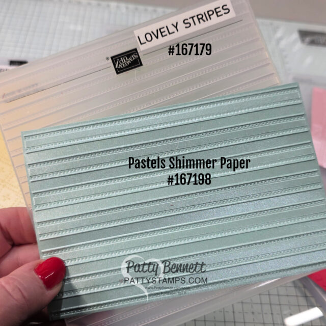 A hand holds a light blue, shimmery embossed paper labeled "Pastels Shimmer Paper #167198" over a striped embossing folder labeled "LOVELY STRIPES #167179" on a craft table. The website "PattyStamps.com" appears on the blue paper. To read more, visit Patty Stamps (pattystamps.com)