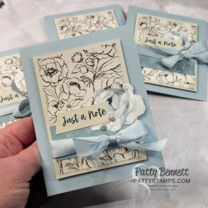A hand holds a light blue greeting card decorated with a floral sketch background, a blue ribbon tied in a bow, a layered paper flower, and a tag that reads "Just a Note." Similar cards are visible on a marble surface in the background. To read more, visit Patty Stamps (pattystamps.com)