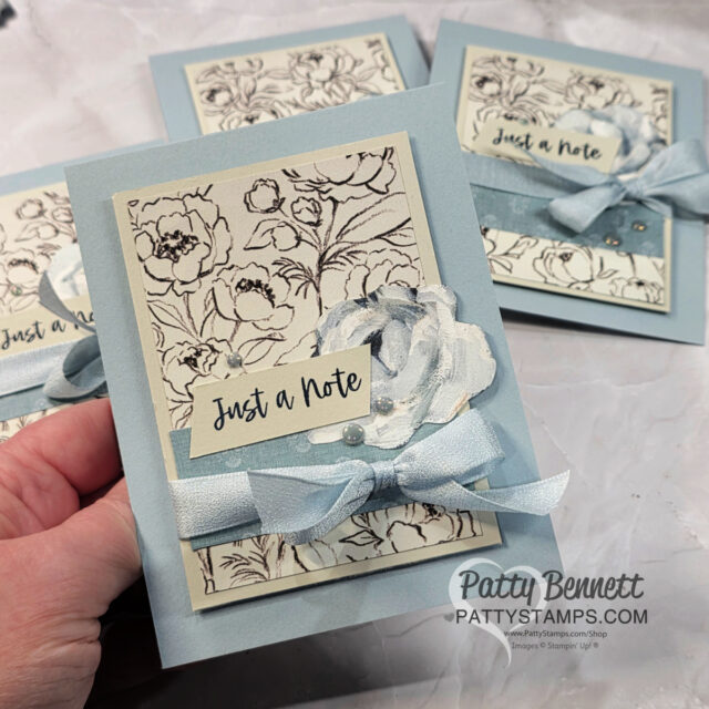 Cloud Cover Card idea. A hand holds a light blue greeting card decorated with a floral sketch background, a blue ribbon tied in a bow, a layered paper flower, and a tag that reads "Just a Note." Similar cards are visible on a marble surface in the background. To read more, visit Patty Stamps (pattystamps.com)
