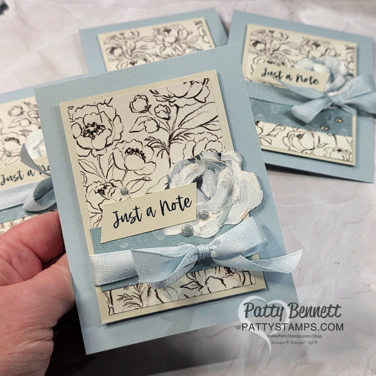 A hand holds a light blue greeting card decorated with a floral sketch background, a blue ribbon tied in a bow, a layered paper flower, and a tag that reads "Just a Note." Similar cards are visible on a marble surface in the background. To read more, visit Patty Stamps (pattystamps.com)