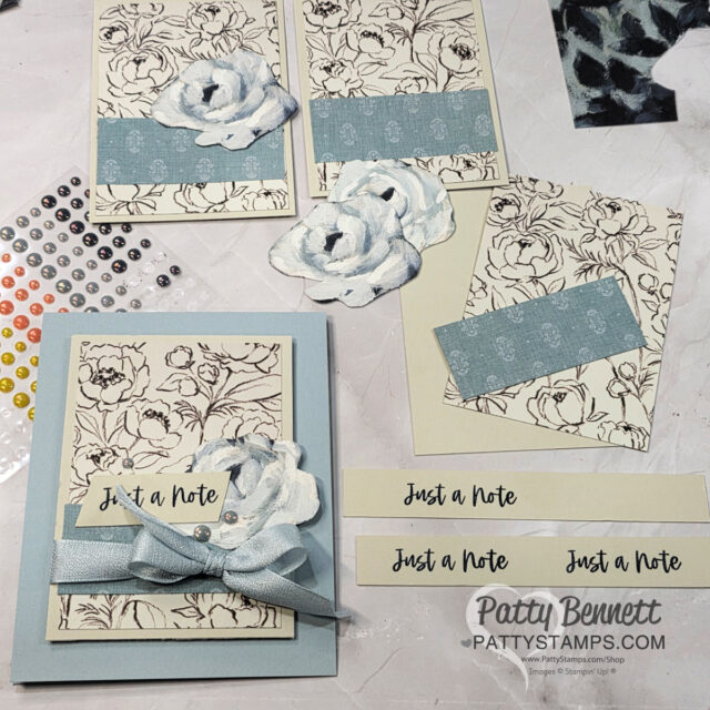 A crafting workspace displays handmade "Just a Note" cards. The cards feature floral line art, blue textured paper strips, 3D white flowers, and ribbon. Adhesive gems, a stencil, and paper scraps are nearby. The watermark reads "Patty Bennett pattystamps.com. To read more, visit Patty Stamps (pattystamps.com)