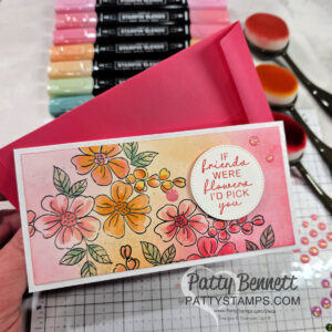 A hand holds a floral greeting card with pink, orange, and yellow flowers and the message “If friends were flowers I’d pick you.” The card is on a pink envelope, surrounded by pastel markers, blending brushes, and decorative gems on a grid mat. To read more, visit Patty Stamps (pattystamps.com)