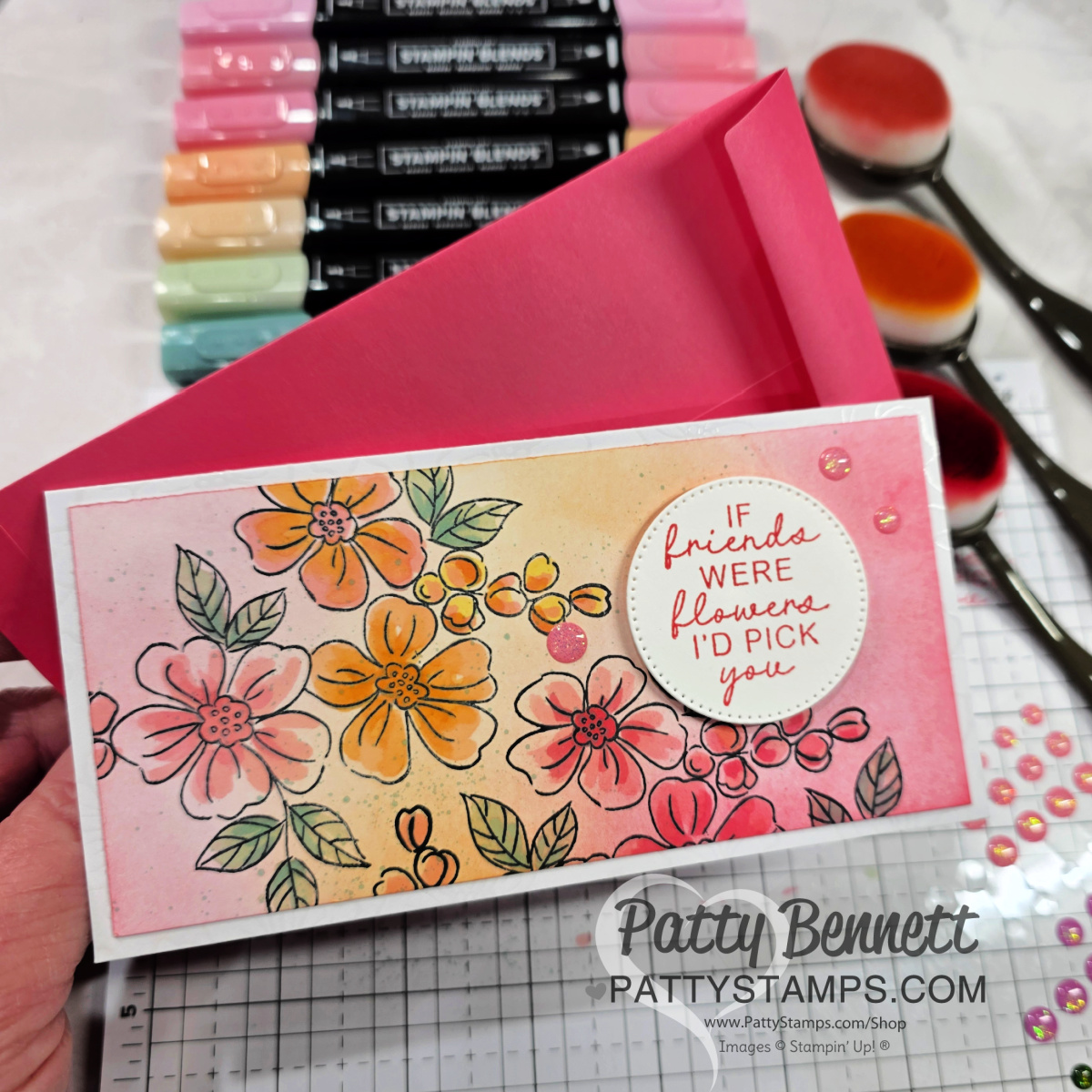 A hand holds a floral greeting card with pink, orange, and yellow flowers and the message “If friends were flowers I’d pick you.” The card is on a pink envelope, surrounded by pastel markers, blending brushes, and decorative gems on a grid mat. To read more, visit Patty Stamps (pattystamps.com)