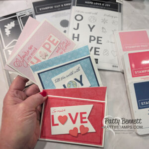 A hand holds four handmade greeting cards, each with layered paper designs and words like “HOPE,” “JOY,” and “LOVE.” In the background, there are stamp sets, craft dies, ink pads, and crafting tools on a white surface. The cards are pink, blue, and red. To read more, visit Patty Stamps (pattystamps.com)