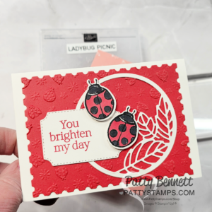 A handmade greeting card features two black and red ladybugs on a red embossed background with strawberries. A white tag reads “You brighten my day.” Decorative leaves and a stitched border enhance the cheerful, layered design. To read more, visit Patty Stamps (pattystamps.com)