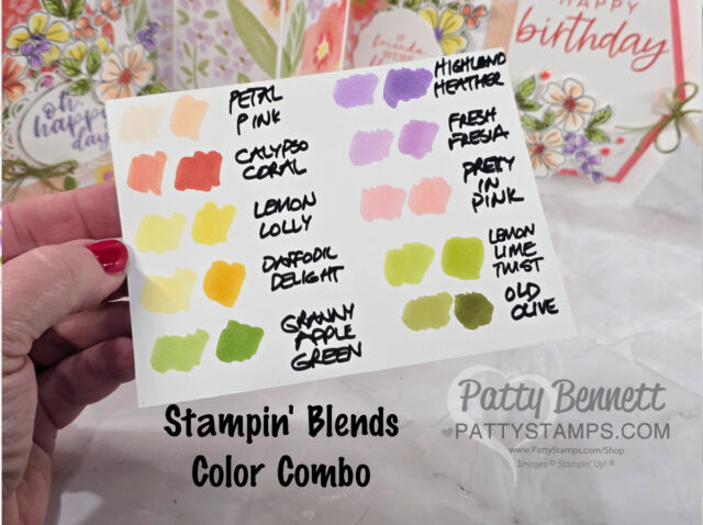 A hand holds a card with twelve colorful ink swatches labeled: Petal Pink, Calypso Coral, Lemon Lolly, Daffodil Delight, Granny Apple Green, Highland Heather, Fresh Freesia, Pretty in Pink, Lemon Lime Twist, and Old Olive. Floral cards are in the background. To read more, visit Patty Stamps (pattystamps.com)