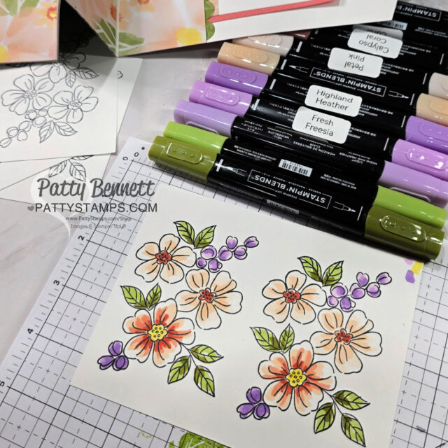 A hand-colored floral card with orange and purple flowers is placed on a grid mat. Stampin’ Blends markers in green, purple, and neutral shades lie above the card. Black and white stamped flower outlines and floral papers are also visible. "Patty Bennett PATTYSTAMPS.COM" is watermarked. To read more, visit Patty Stamps (pattystamps.com)