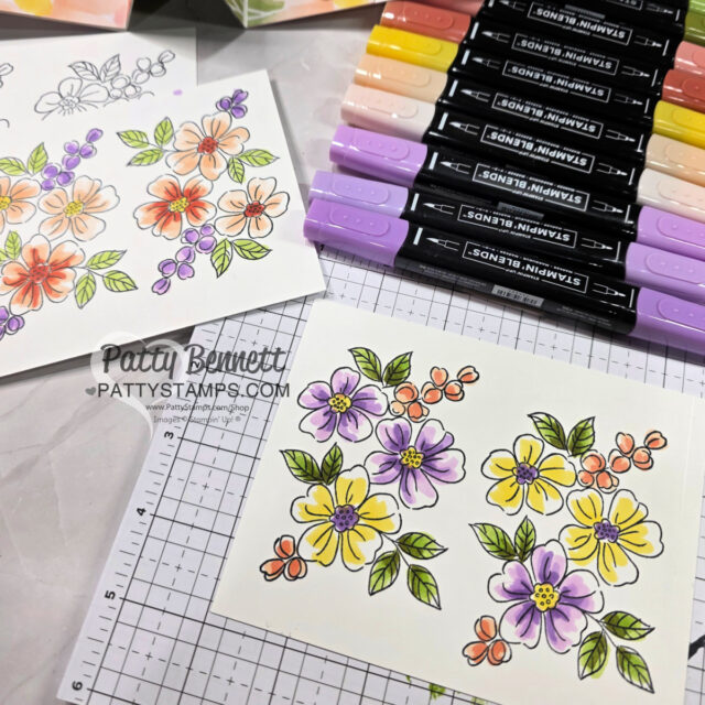 Two white cards lie on a grid mat, each decorated with hand-colored floral designs in orange, purple, and yellow tones. Several Stampin’ Blends markers in pastel colors are arranged nearby. One card displays the watermark “Patty Bennett PATTTYSTAMPS.COM.”. To read more, visit Patty Stamps (pattystamps.com)