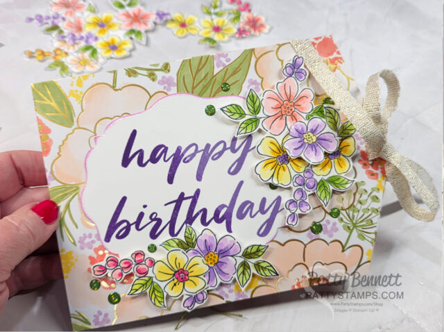 A hand holds a floral-themed birthday card with “happy birthday” in bold purple script. The card is decorated with colorful, hand-drawn flowers, green leaves, and a cream ribbon tied in a bow on the right edge. A few more flowers are blurred in the background. To read more, visit Patty Stamps (pattystamps.com)
