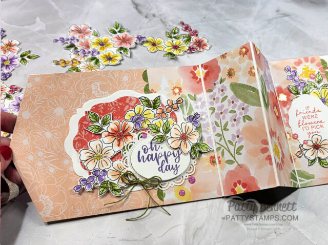 An open handmade card with floral patterns and layered, colorful paper flowers. The central text reads “oh, happy day.” Inside, another floral cluster frames the words “If friends were flowers, I’d pick you.” More flower cutouts are scattered nearby. To read more, visit Patty Stamps (pattystamps.com)