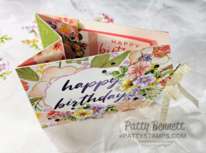 A handmade floral card with “happy birthday” written on the front in purple script. The card has colorful flowers, layered paper, and a gold ribbon tied to the side. The inside shows another “happy birthday” message and more floral designs. To read more, visit Patty Stamps (pattystamps.com)