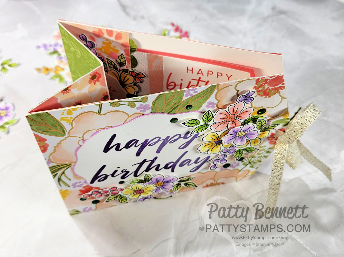 A handmade floral card with “happy birthday” written on the front in purple script. The card has colorful flowers, layered paper, and a gold ribbon tied to the side. The inside shows another “happy birthday” message and more floral designs. To read more, visit Patty Stamps (pattystamps.com)