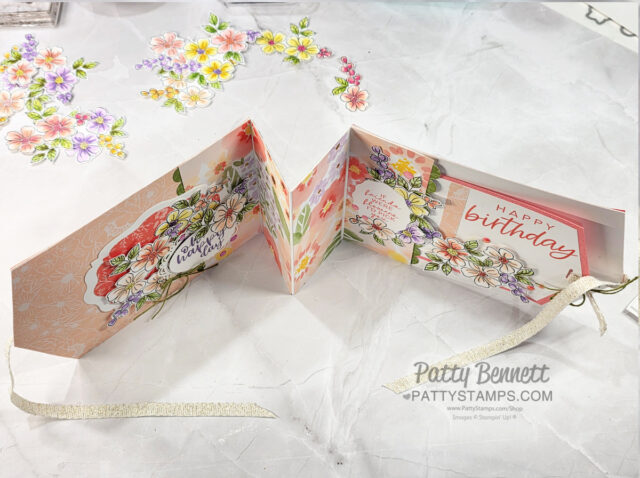A handmade, folded birthday card is laid open on a white surface. The card features pastel floral designs, "Happy Birthday" text, and layered embellishments. Loose floral cutouts and a pale ribbon are nearby. "Patty Bennett" and her website are watermarked. To read more, visit Patty Stamps (pattystamps.com)
