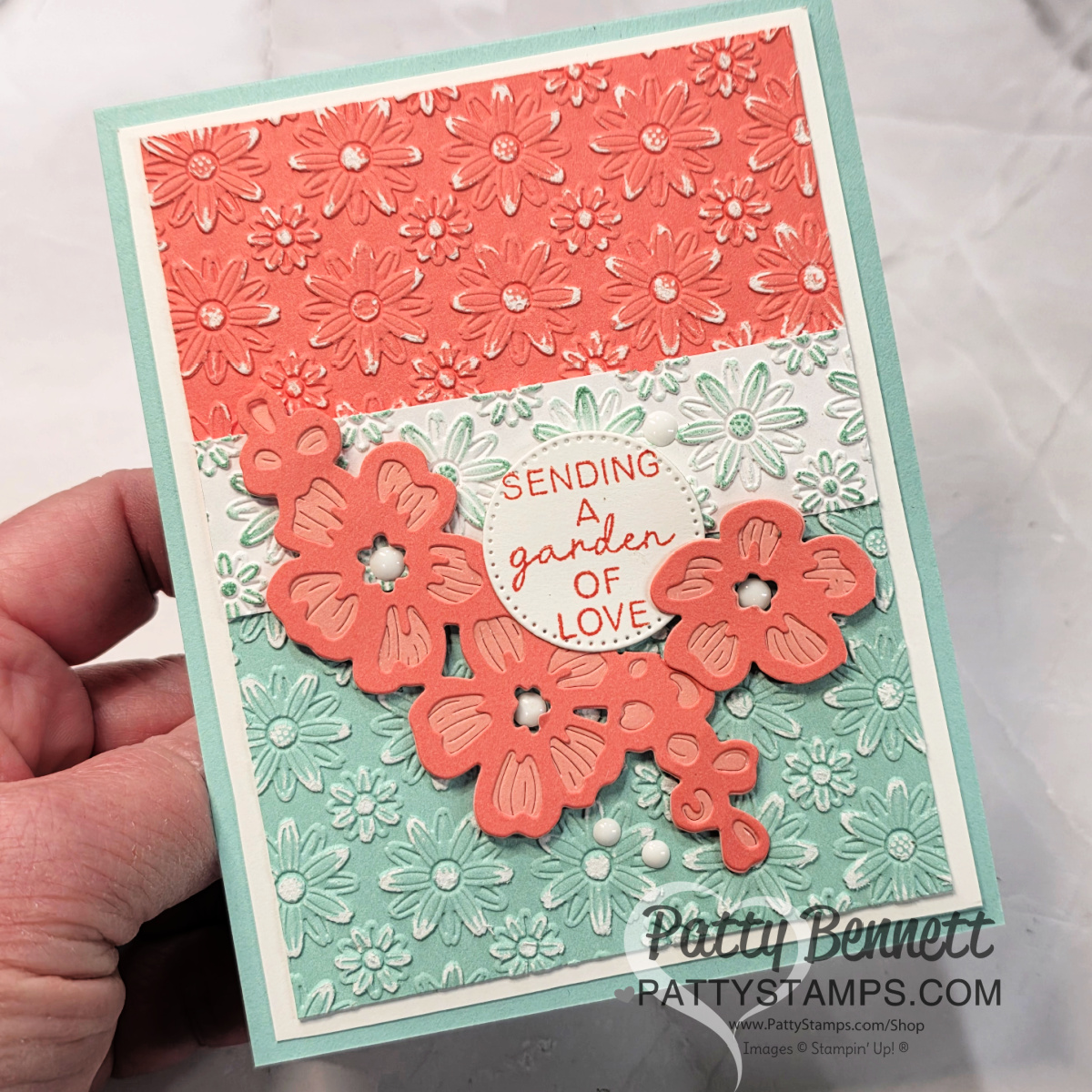 A handmade greeting card with embossed coral and mint green floral designs, coral die-cut flowers, and a central white label reading "SENDING A garden OF LOVE." A hand holds the card over a white background. Watermark: Patty Bennett pattystamps.com. To read more, visit Patty Stamps (pattystamps.com)