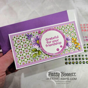 A hand holds a handmade card with floral designs, a purple and white geometric background, and the message “Grateful for your friendship” in a circle. The card is paired with a purple envelope. Colorful adhesive gems are visible nearby. To read more, visit Patty Stamps (pattystamps.com)