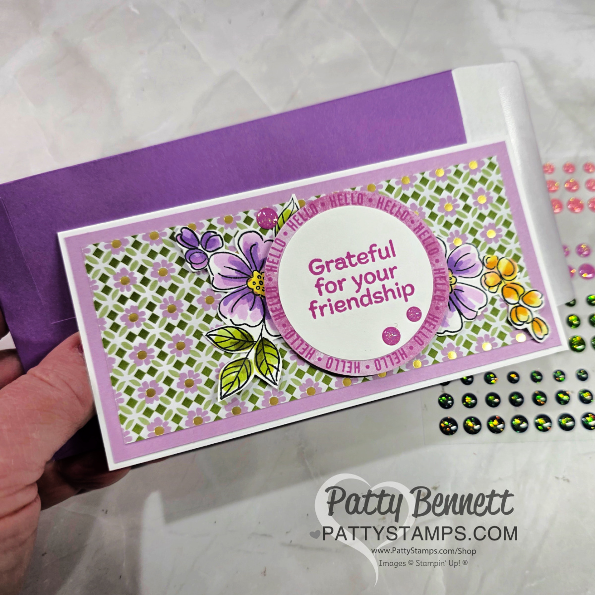 A hand holds a handmade card with floral designs, a purple and white geometric background, and the message “Grateful for your friendship” in a circle. The card is paired with a purple envelope. Colorful adhesive gems are visible nearby. To read more, visit Patty Stamps (pattystamps.com)