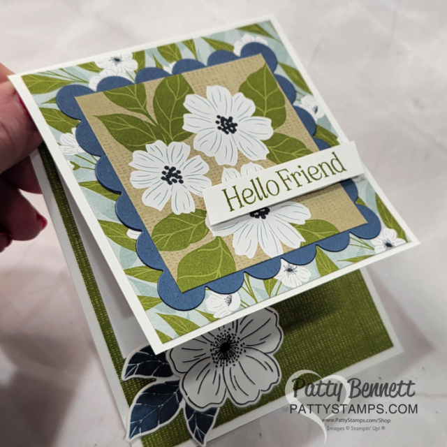 A handmade greeting card with a floral design and “Hello Friend” sentiment. The card features layered papers with white flowers and green leaves, scalloped blue borders, and a large flower embellishment on the bottom left. “Patty Bennett” watermark is at the bottom right. To read more, visit Patty Stamps (pattystamps.com)