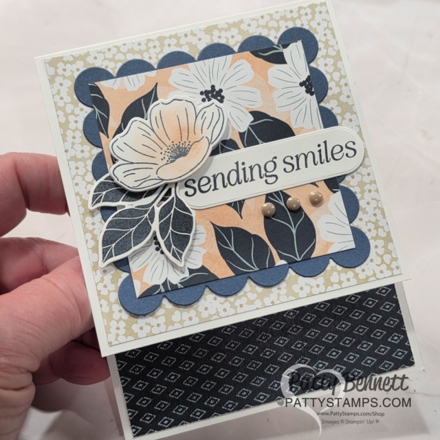 A handmade greeting card with layered floral patterns in cream, navy, and peach, featuring large illustrated flowers and leaves. The front displays the sentiment “sending smiles” and three beige dots. A hand holds the card, which is partially open to reveal a patterned interior. To read more, visit Patty Stamps (pattystamps.com)