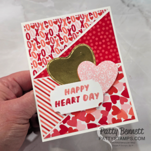 A handmade Valentine’s Day card with red, pink, and white patterned sections, featuring hearts and "XOXO" motifs. It is decorated with gold and pink hearts and a white label reading "HAPPY HEART DAY." A hand holds the card on a marble background. To read more, visit Patty Stamps (pattystamps.com)