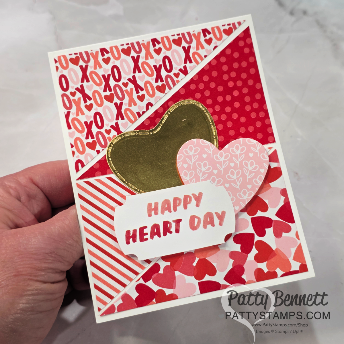 A handmade Valentine’s Day card with red, pink, and white patterned sections, featuring hearts and "XOXO" motifs. It is decorated with gold and pink hearts and a white label reading "HAPPY HEART DAY." A hand holds the card on a marble background. To read more, visit Patty Stamps (pattystamps.com)