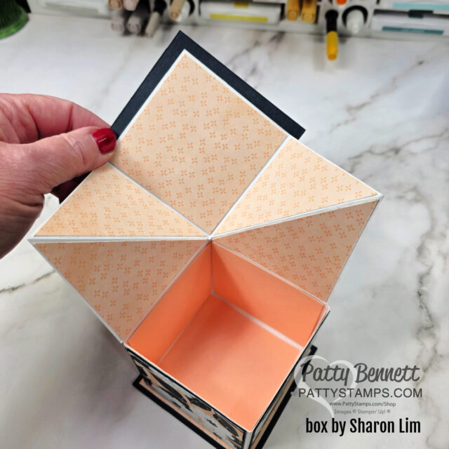 Pop Up Box and video. A hand opens an origami-style box with a peach and cream patterned lid divided into triangle sections. The box interior is peach-colored. Text on the image credits Patty Bennett at PattyStamps.com and notes the box is made by Sharon Lim. To read more, visit Patty Stamps (pattystamps.com)