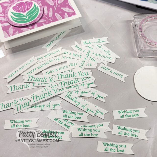 A table displays numerous small white paper banners with green and gray stamped phrases like “Thank You,” “Wishing you all the best,” “Happy Birthday,” and “Just a Note.” A handmade card with a pink flower and circle pattern sits in the background. To read more, visit Patty Stamps (pattystamps.com)
