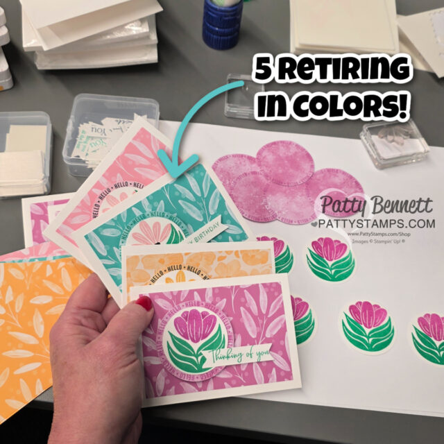 A hand holds five floral greeting cards in pink, orange, yellow, green, and purple. An arrow labeled "5 Retiring In Colors!" points to the cards. Craft supplies, ink pads, and stamped flower cutouts are on the table in the background. To read more, visit Patty Stamps (pattystamps.com)