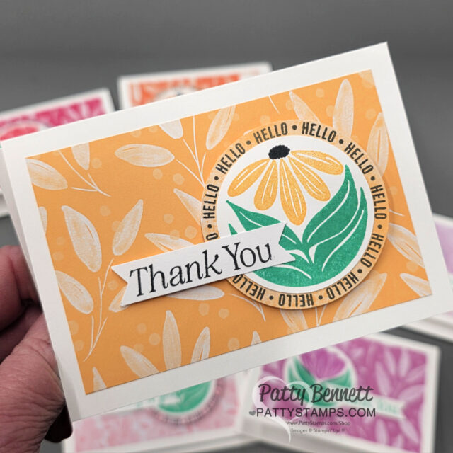 A hand holds a greeting card with an orange leafy background, a daisy illustration in a circle labeled "HELLO" repeatedly, and a white banner reading "Thank You." The card is signed "Patty Bennett, Pattystamps.com." More cards are blurred in the background. To read more, visit Patty Stamps (pattystamps.com)