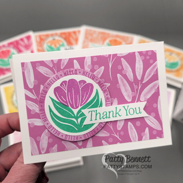 A hand holds a greeting card with a pink and purple floral design. The card features a purple circular frame with "HELLO" repeated, a tulip illustration, and a green "Thank You" banner. Similar cards in orange, pink, and teal are blurred in the background. To read more, visit Patty Stamps (pattystamps.com)