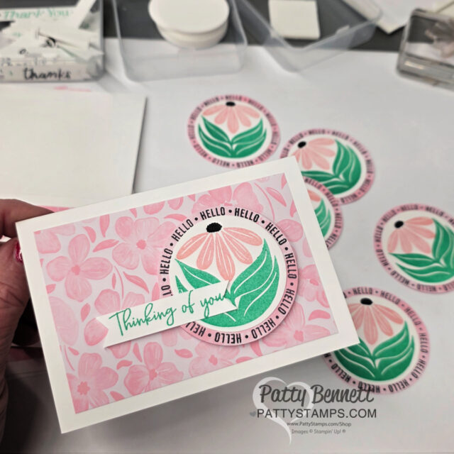 A hand holds a greeting card decorated with pink floral patterns, a circular design with a pink and black daisy, green leaves, and repeated “HELLO” text around it. The card reads “Thinking of you.” Several similar circular floral designs are on the table nearby. To read more, visit Patty Stamps (pattystamps.com)