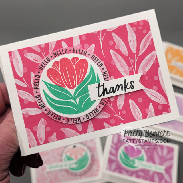 A hand holds a greeting card with a red and pink floral design on a pink background. The card features a circular "HELLO" border around a stylized flower and a white banner with "thanks" in black script. Other similar cards are blurred in the background. To read more, visit Patty Stamps (pattystamps.com)