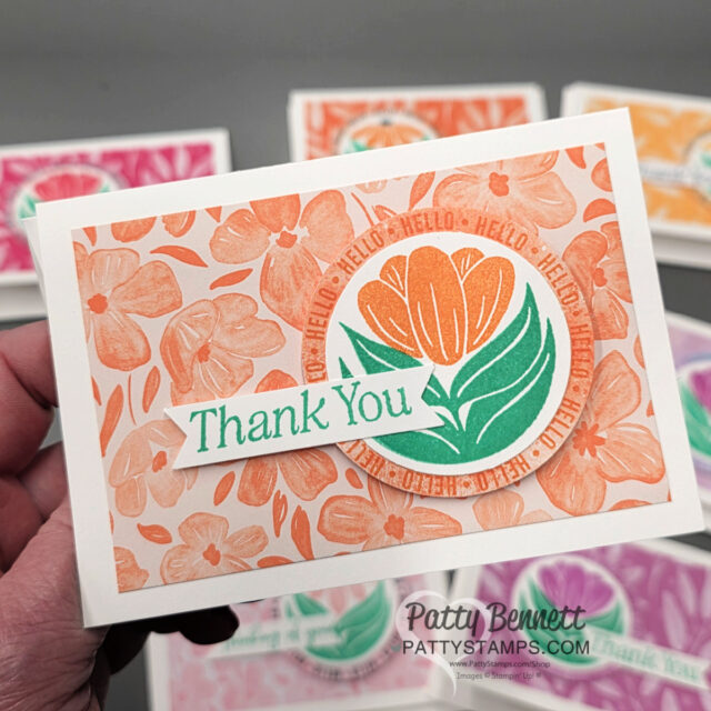 A hand holds a card with peach and orange floral patterns, a stylized orange flower in a green circle, and repeated "HELLO" text. A diagonal banner with "Thank You" in green is on top. Similar cards in other colors are blurred in the background. To read more, visit Patty Stamps (pattystamps.com)